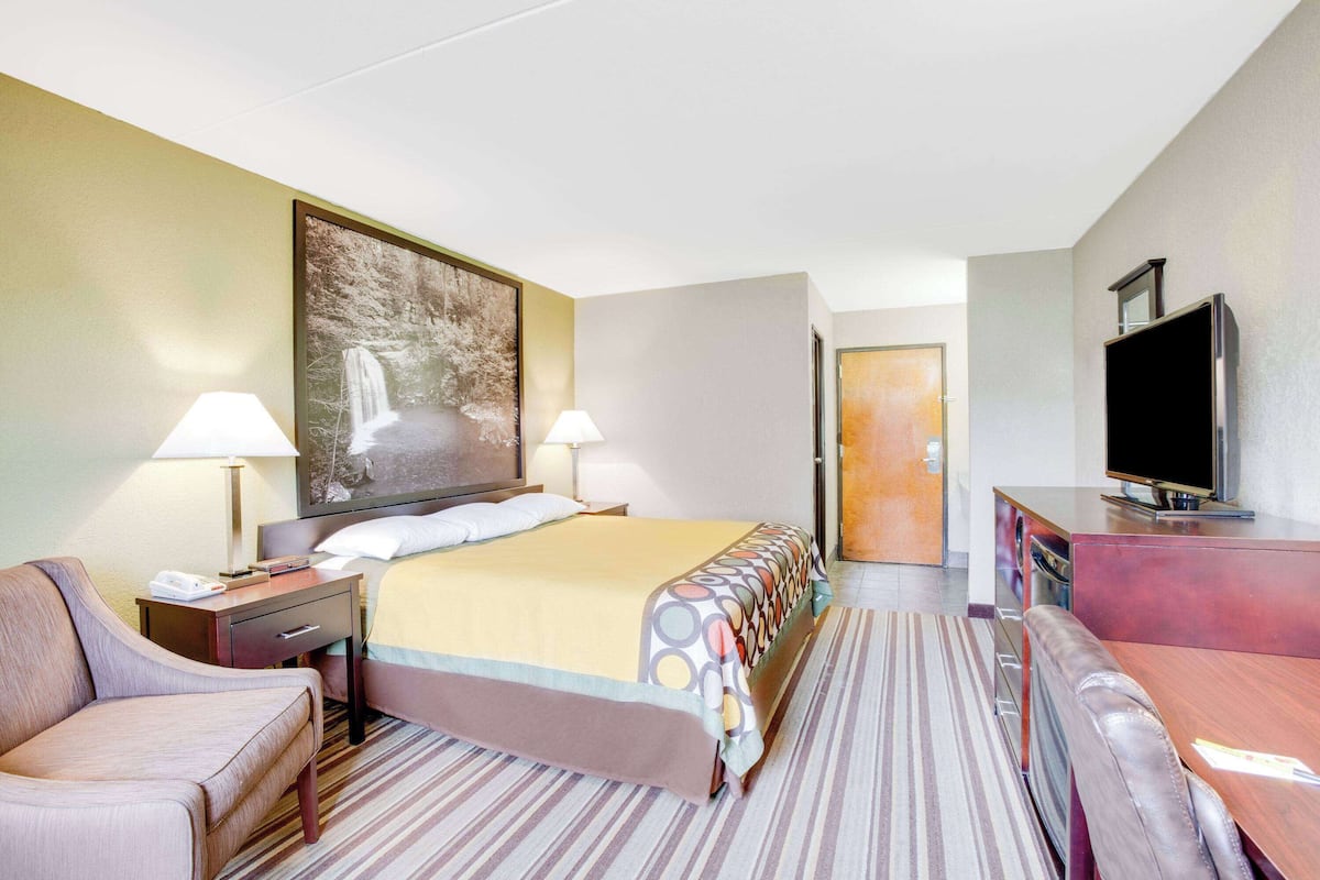 A hotel room with a bed, two bedside lamps, a television, a desk, a chair, and a painting on the wall.