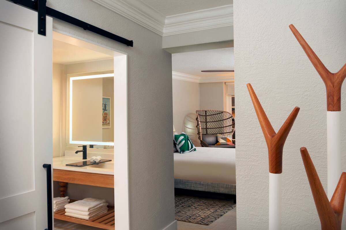 A hotel room with a bathroom featuring a sink, mirror, and towel rack. There is a bed with pillows and a green bag on it.