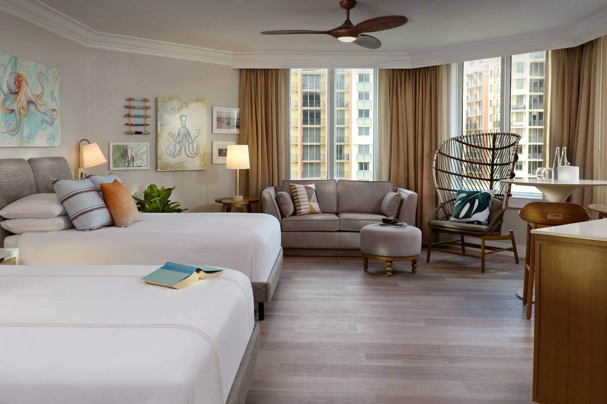 A hotel room with two beds, a sofa, a chair, and a dining table. There are large windows with curtains, artwork on the walls, and a ceiling fan.