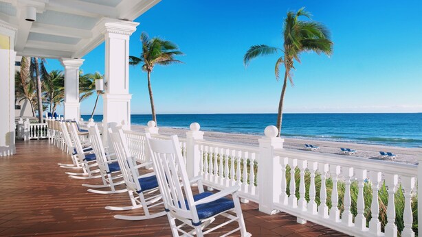 A beachside terrace with white rocking chairs and a white balustrade overlooking the ocean. - Pelican Grand Beach Resort - A Noble House Resort (Fort Lauderdale)