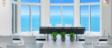 A modern dining area with a view of the sea, featuring a long table with chairs, potted plants, and a bar area.