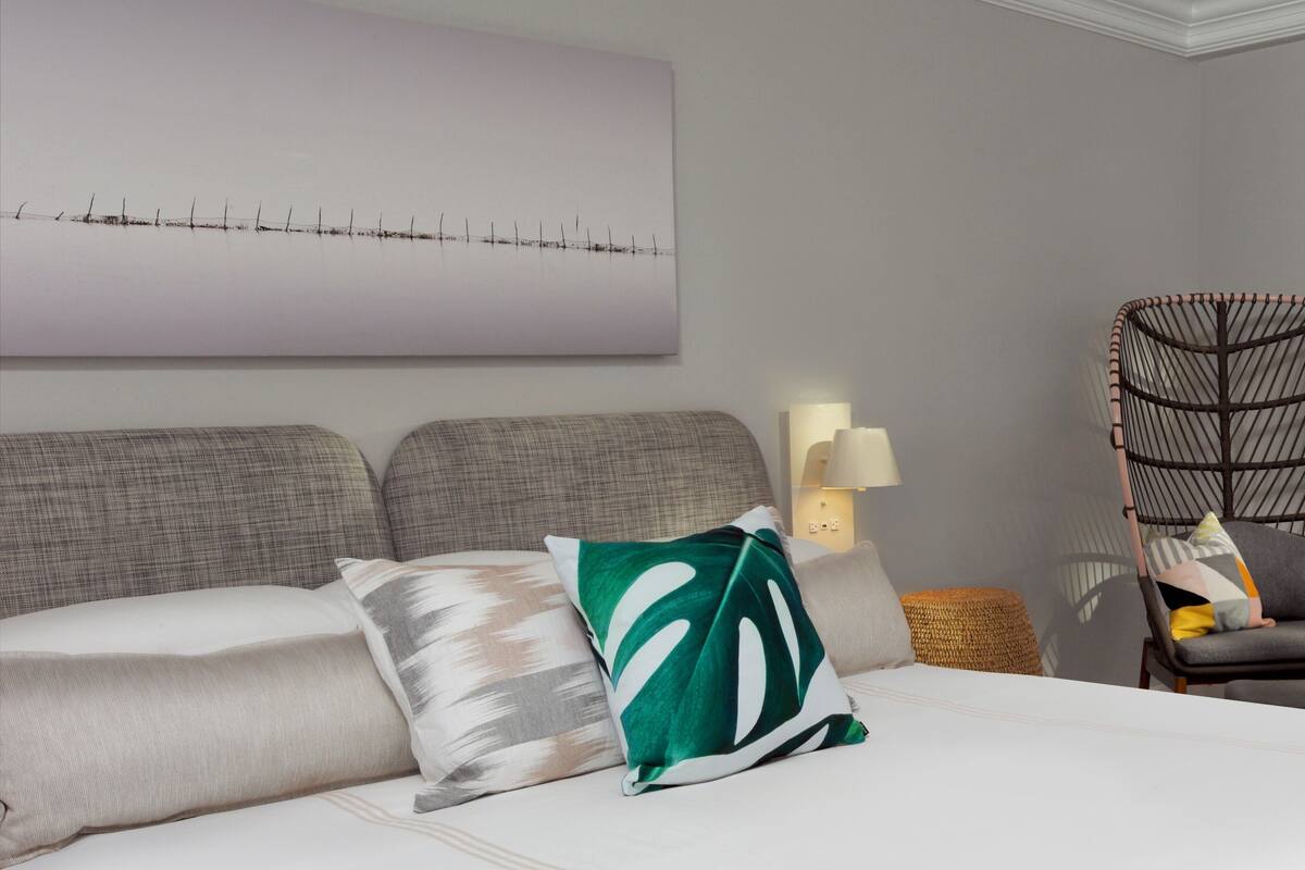 A hotel room with a large bed, a headboard, a wall-mounted artwork, a side table with a lamp, and a wicker chair.