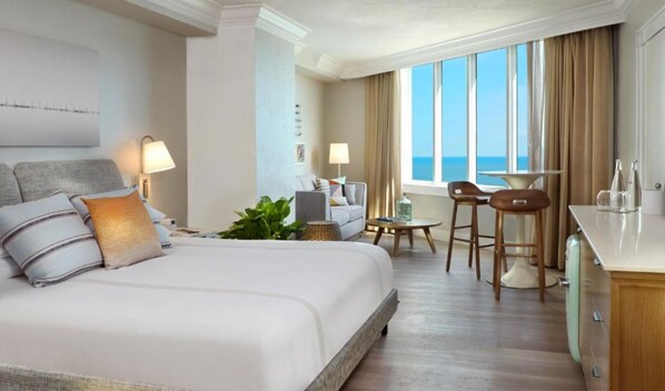 A hotel room with a large bed, a desk, two chairs, a small table, and a view of the ocean.