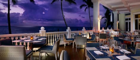 A restaurant with outdoor seating overlooking the ocean, featuring wicker chairs and tables set for dining.