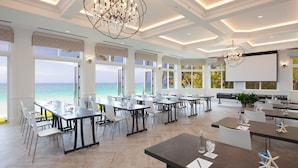 A spacious conference room with large windows overlooking the ocean, equipped with tables, chairs, and a projector screen. - Pelican Grand Beach Resort - A Noble House Resort (Fort Lauderdale)