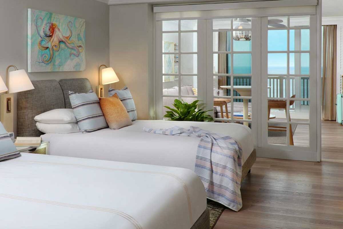 A hotel room with two beds, a painting on the wall, and a balcony with a view.