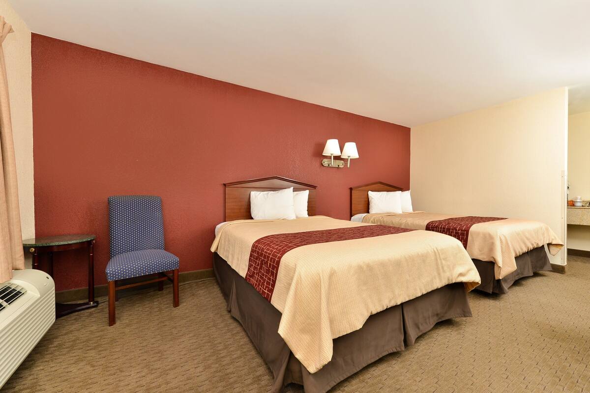 A hotel room with two beds, a chair, a table, and a wall-mounted lamp.