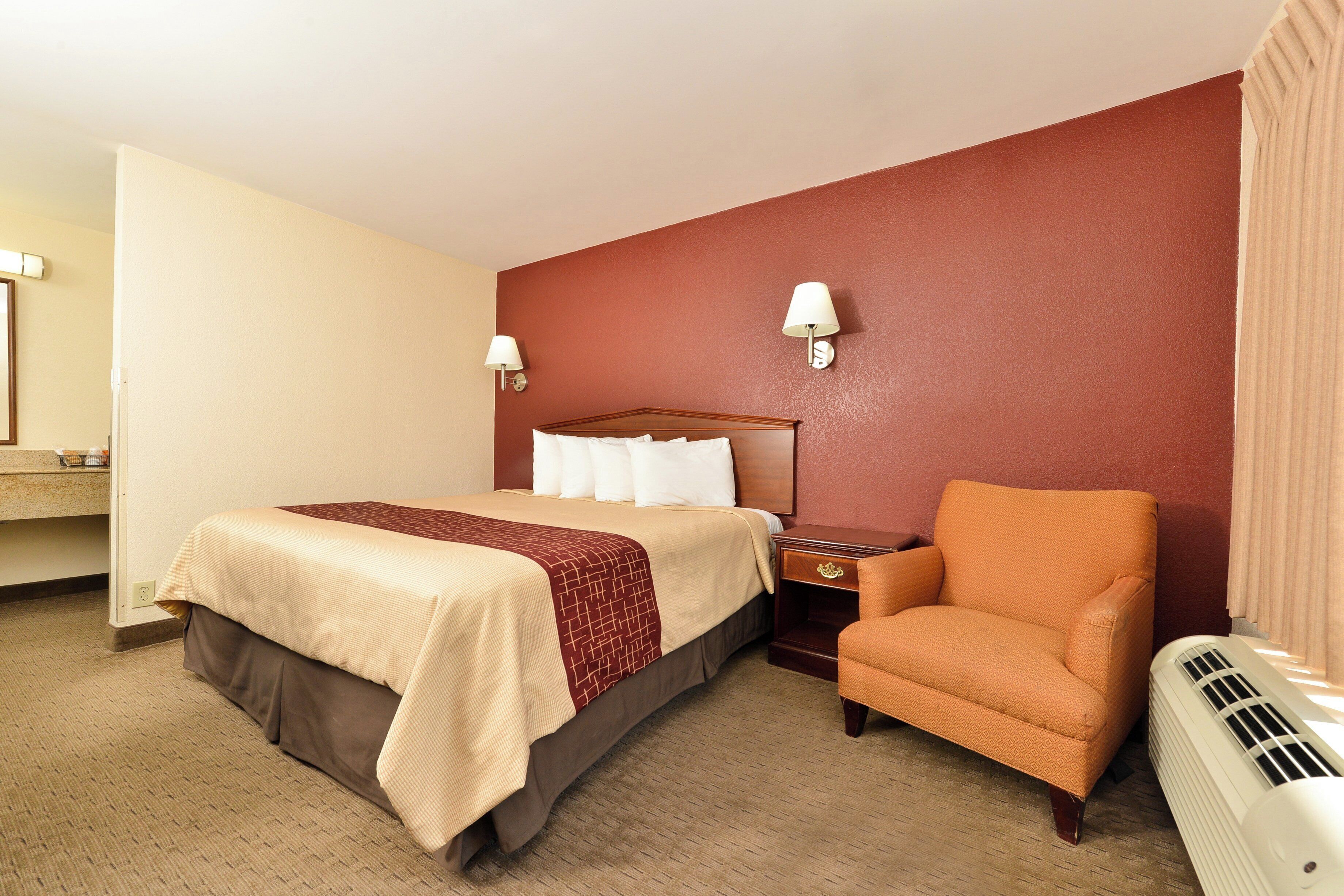 Una habitación de hotel con una cama, una silla, una mesita de noche, una lámpara y un calentador.