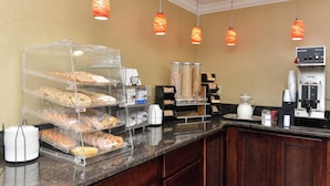 Un'area colazione self-service con una macchina del caffè, una caraffa e diversi dolci e pane esposti in contenitori trasparenti.