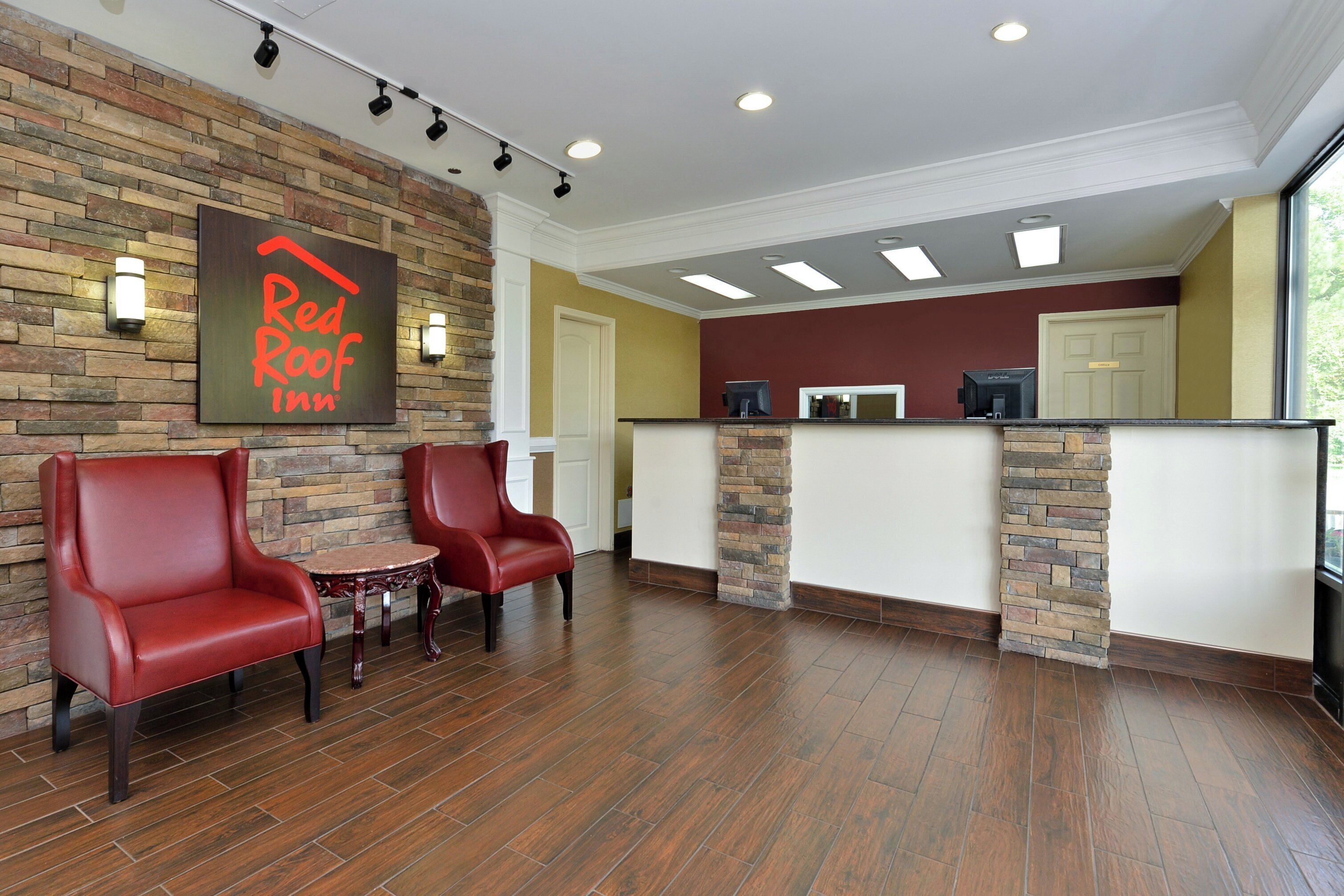 A Red Roof Inn reception area with a stone wall, two red leather chairs, a small table, and a reception desk with a computer.