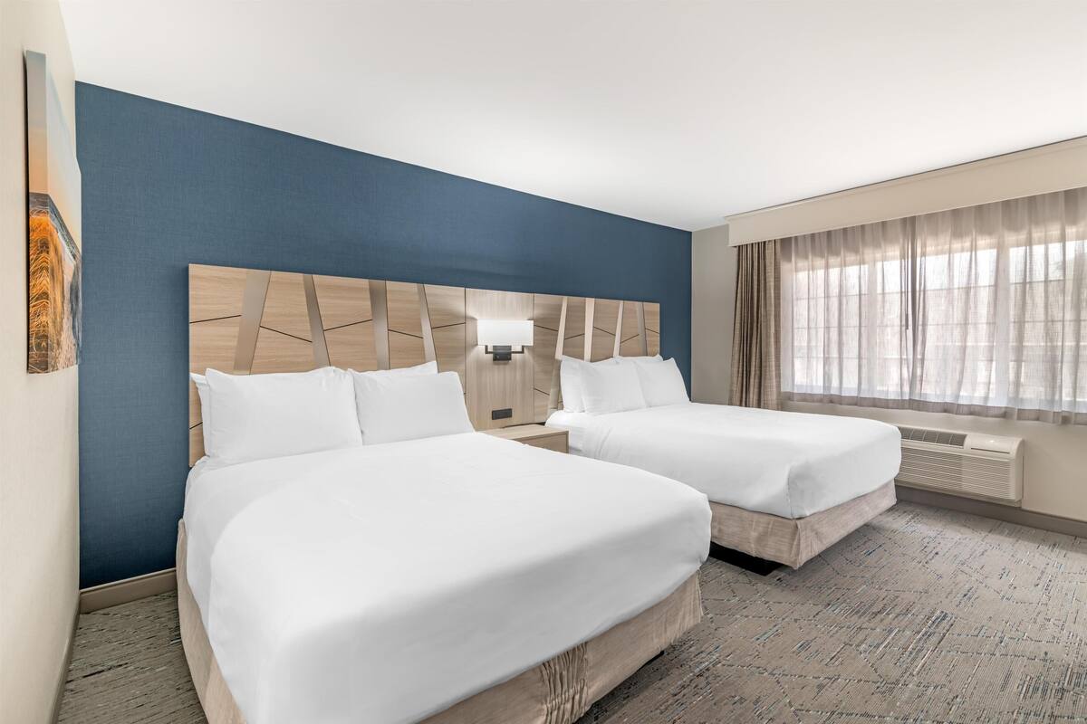 A hotel room with two beds, a large wall-mounted headboard, a window with sheer curtains, and a wall-mounted air conditioning unit.