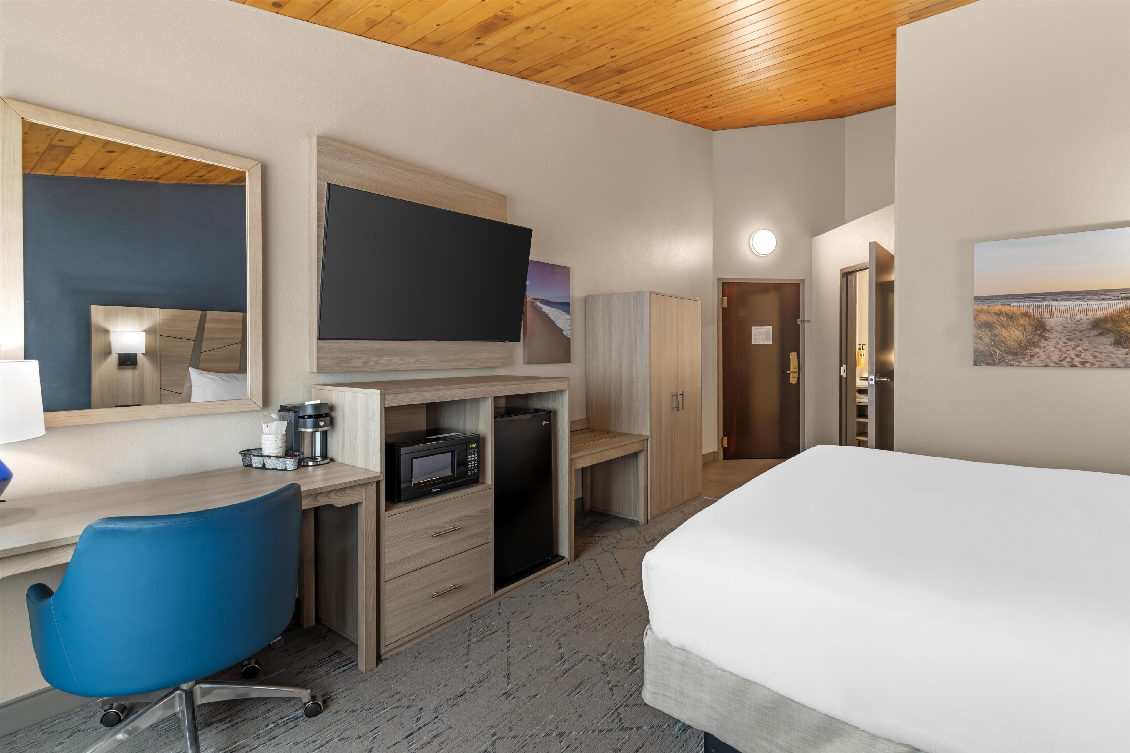 A hotel room with a bed, a desk with a chair, a television, a microwave, a refrigerator, and a door leading to another room.