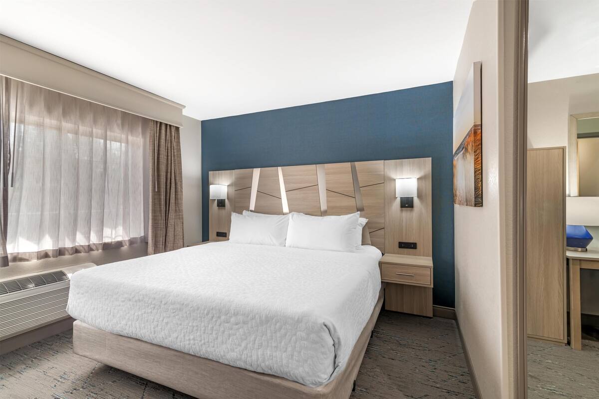 A hotel room with a large bed, a bedside table, a nightstand, a wall-mounted lamp, and a window with curtains.