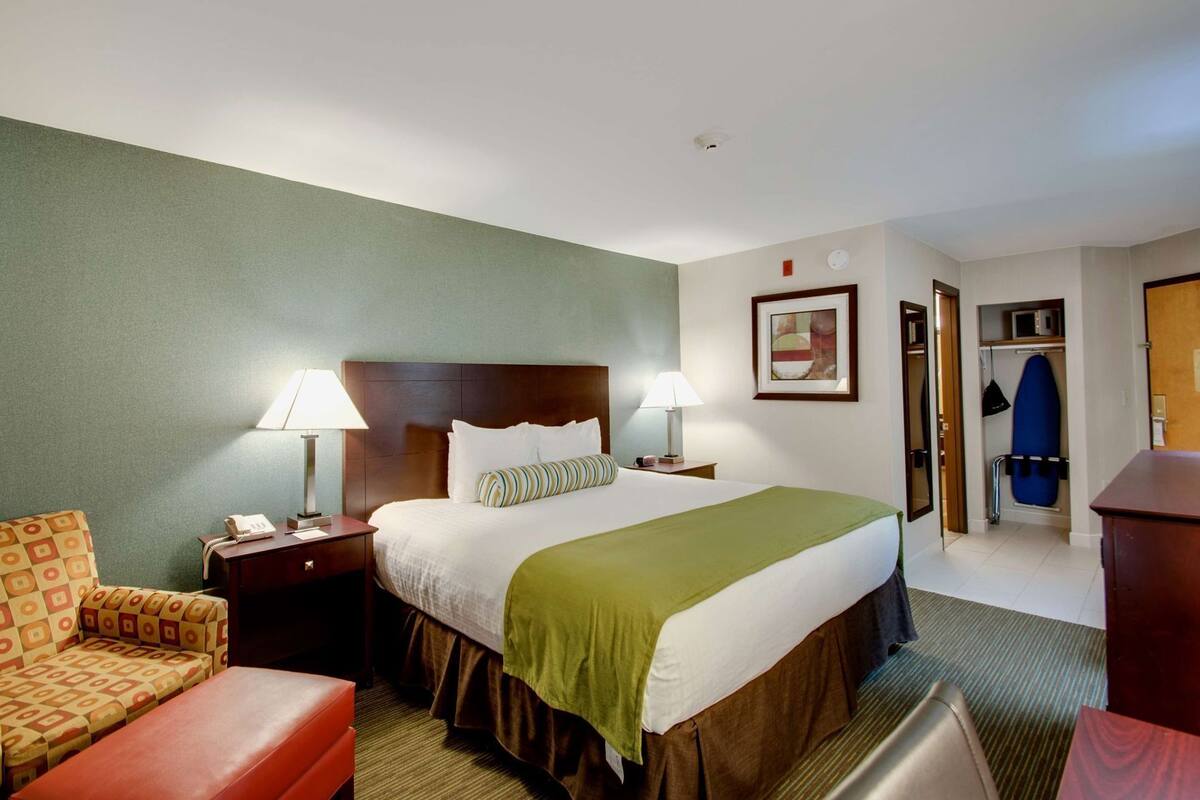 A hotel room with a bed, two bedside lamps, a chair, a desk, a coat rack, and a door leading to a bathroom.