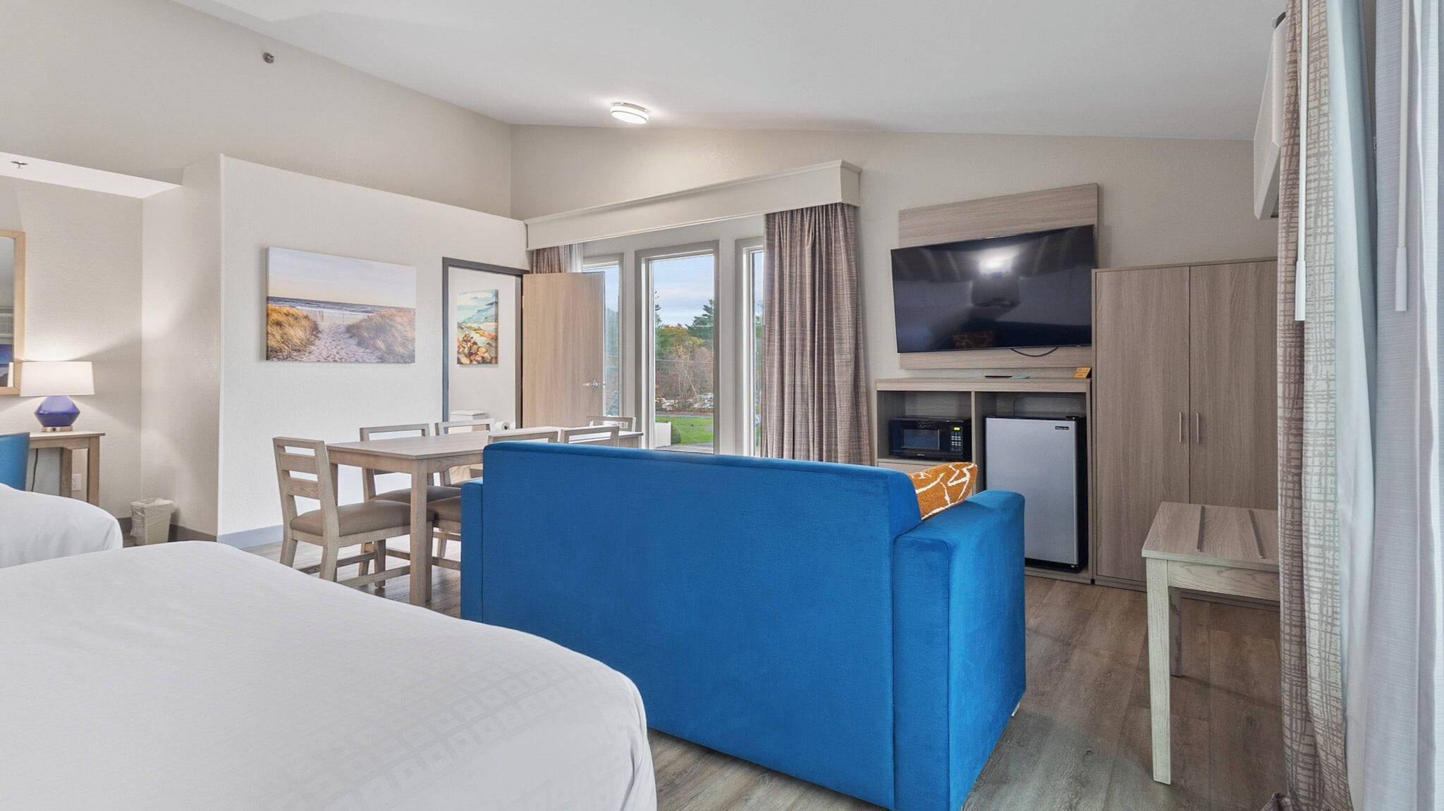 A hotel room with a bed, a blue sofa, a dining table with chairs, a TV, a microwave, and a refrigerator.