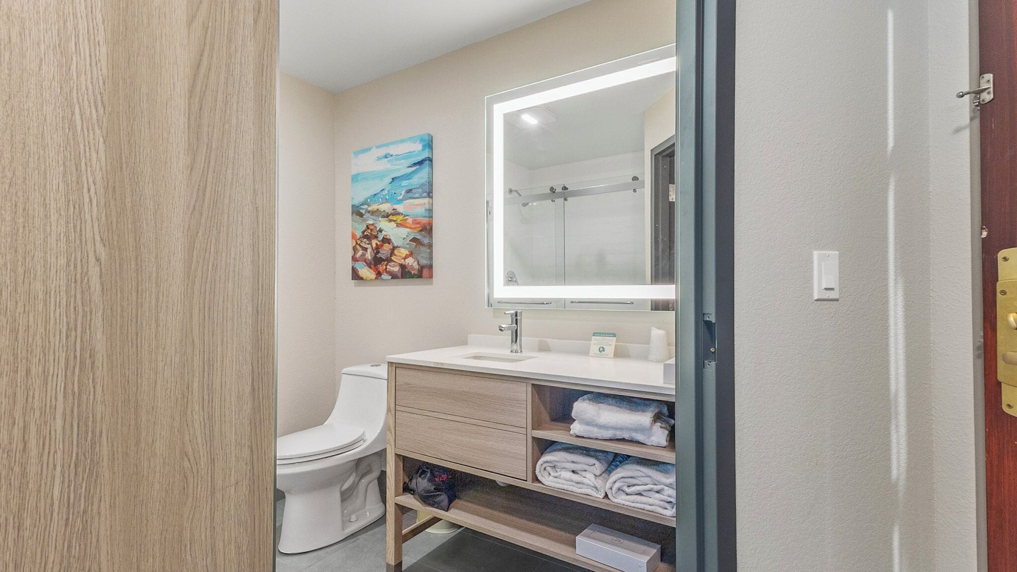 A modern bathroom with a white toilet, a sink with a large mirror, and a shelf with towels.