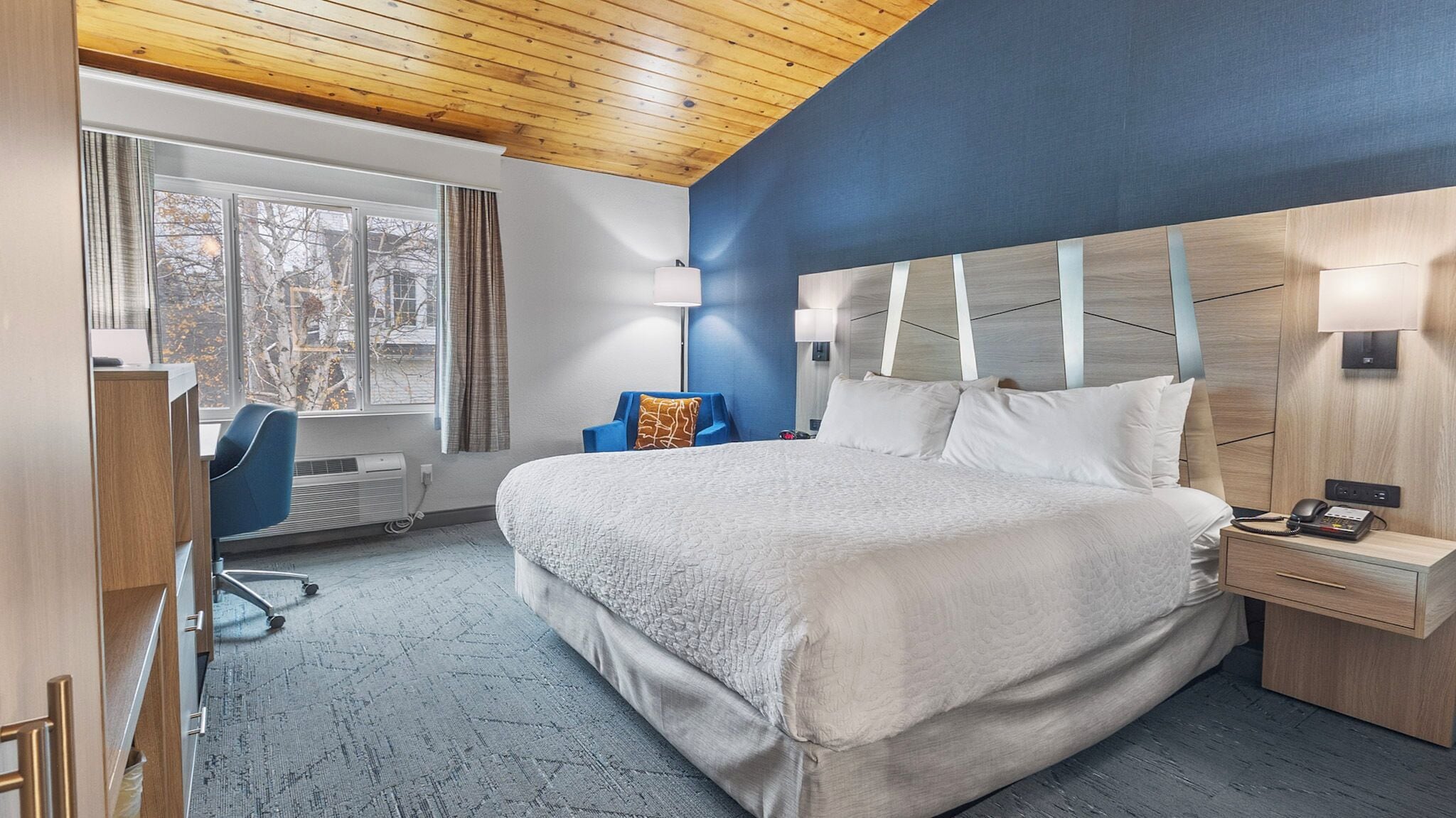 Quarto de hotel com uma cama grande, uma escrivaninha, uma cadeira e uma janela com cortinas.