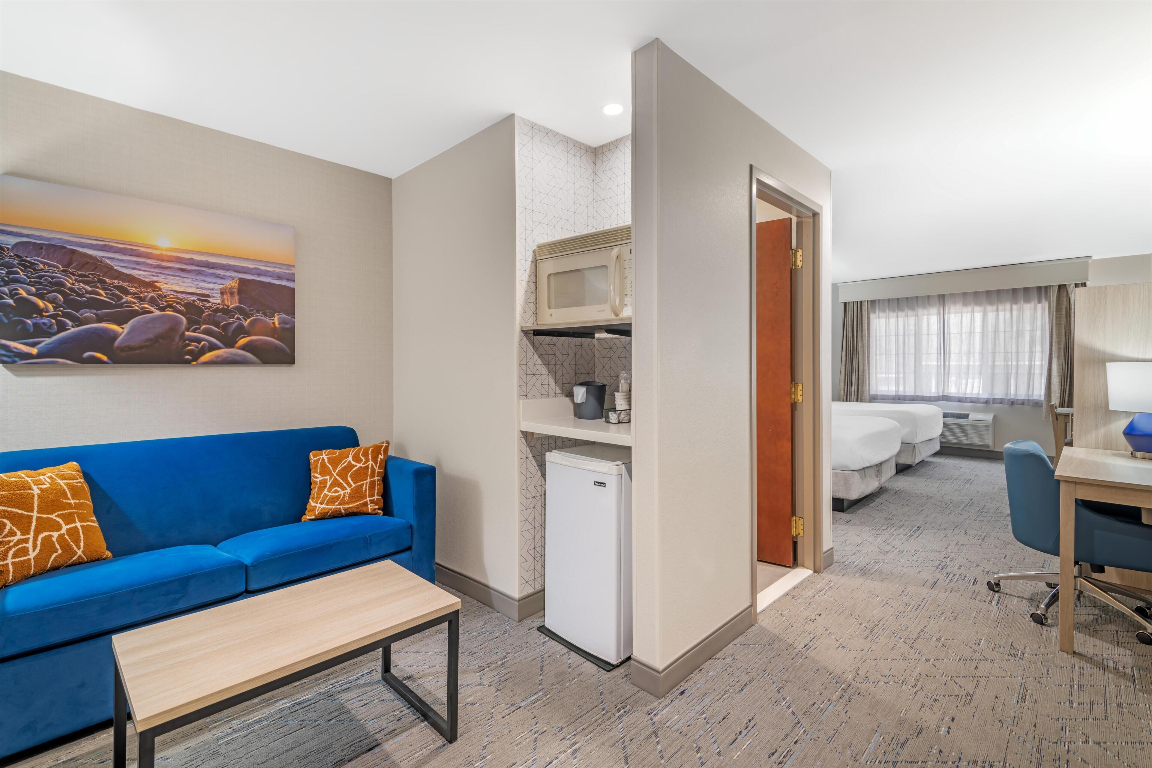 A hotel room with a blue sofa, a small table, a microwave, a mini-fridge, a bed, a desk, and a painting of a beach scene.