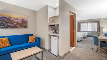 A hotel room with a blue sofa, a small table, a microwave, a mini-fridge, a bed, a desk, and a painting of a beach scene.