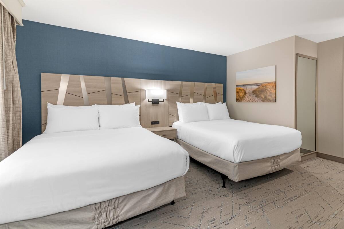 A hotel room with two beds, a headboard with a geometric design, a nightstand with a lamp, a painting on the wall, and a carpeted floor.