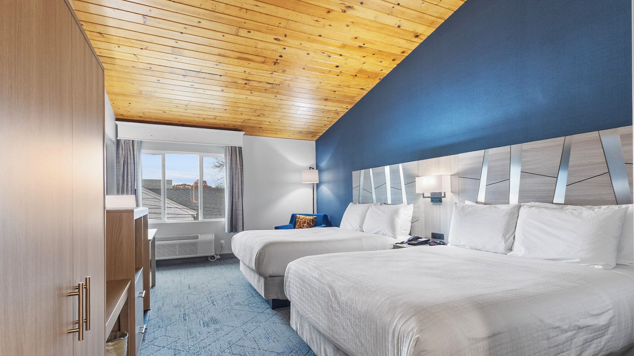 A hotel room with two beds, a wooden ceiling, a blue accent wall, and a window with curtains.