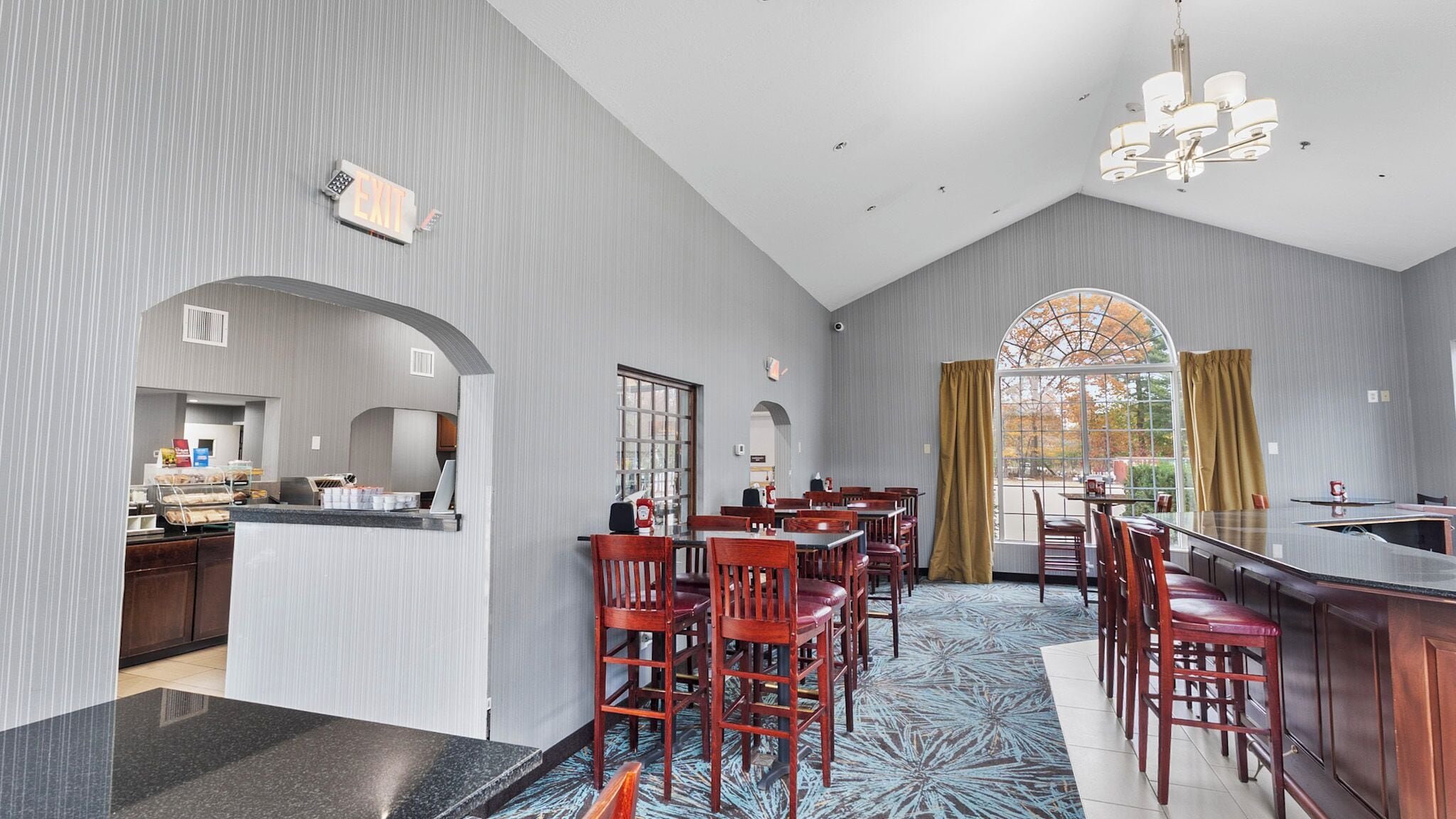 A modern hotel dining area with a bar, high chairs, and a large window with curtains.