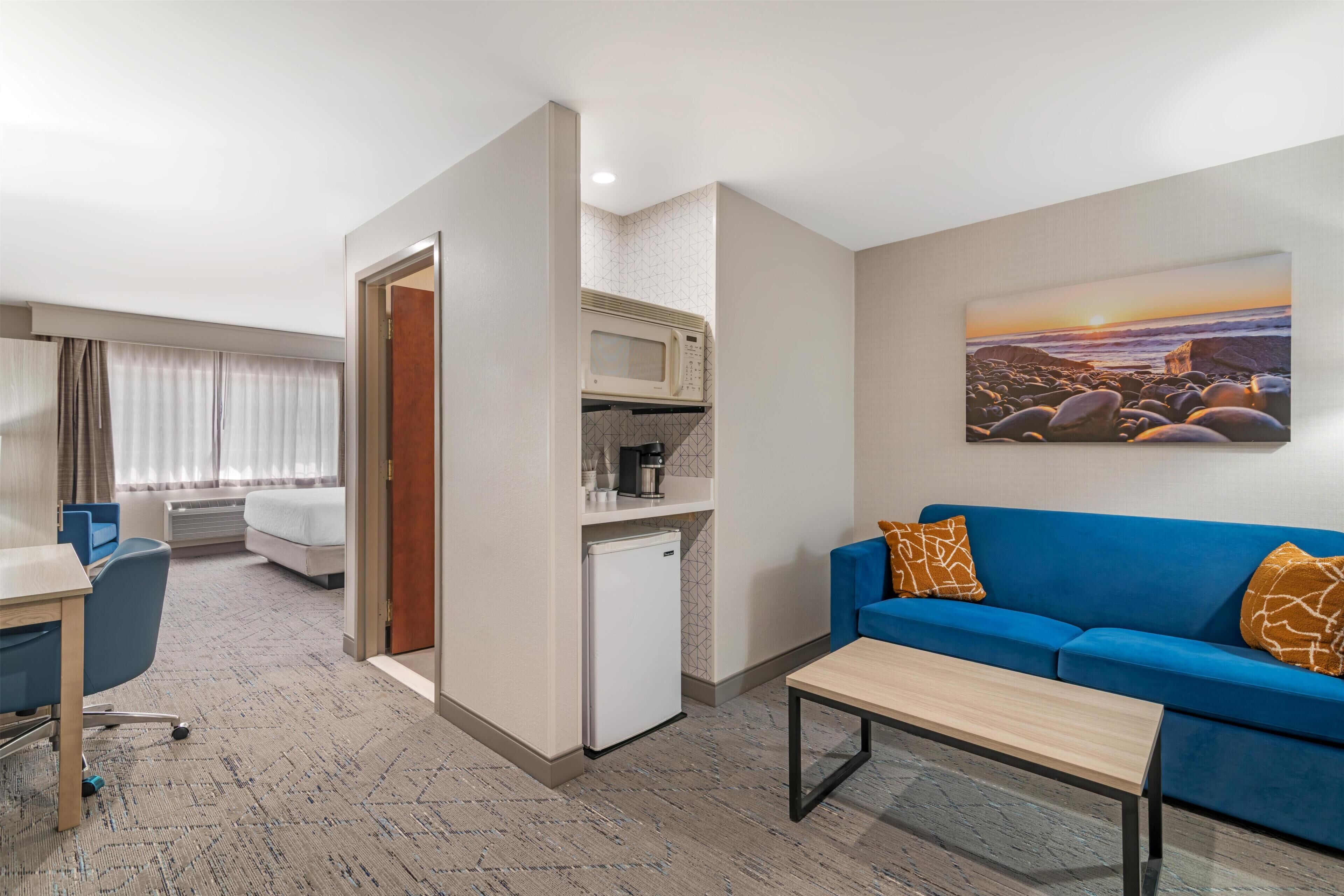 A hotel room with a kitchenette, a blue sofa, a painting of a sunset, and a bed with curtains.