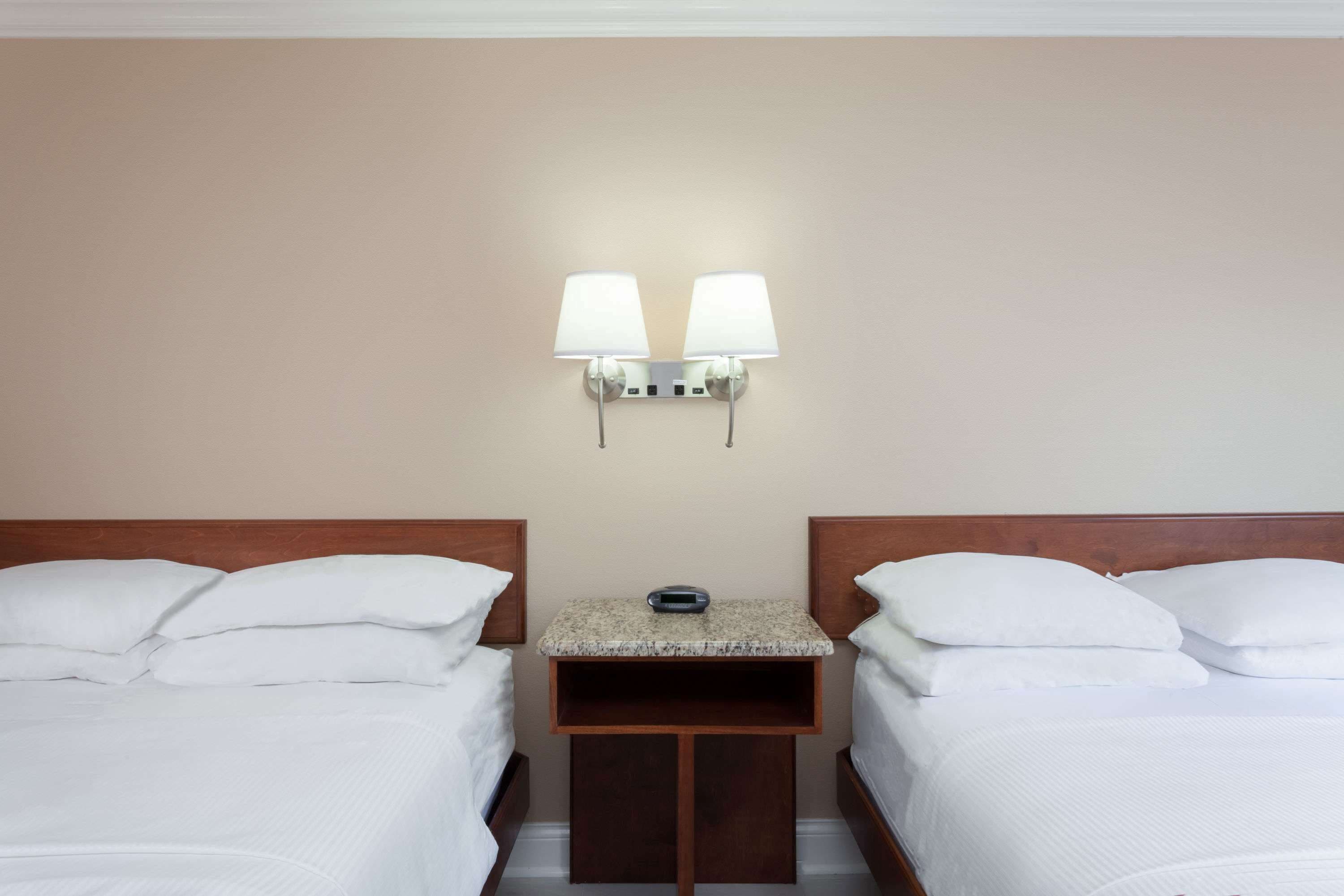 Habitación de hotel con dos camas, una mesita de noche de madera con lámpara y una lámpara de pared.