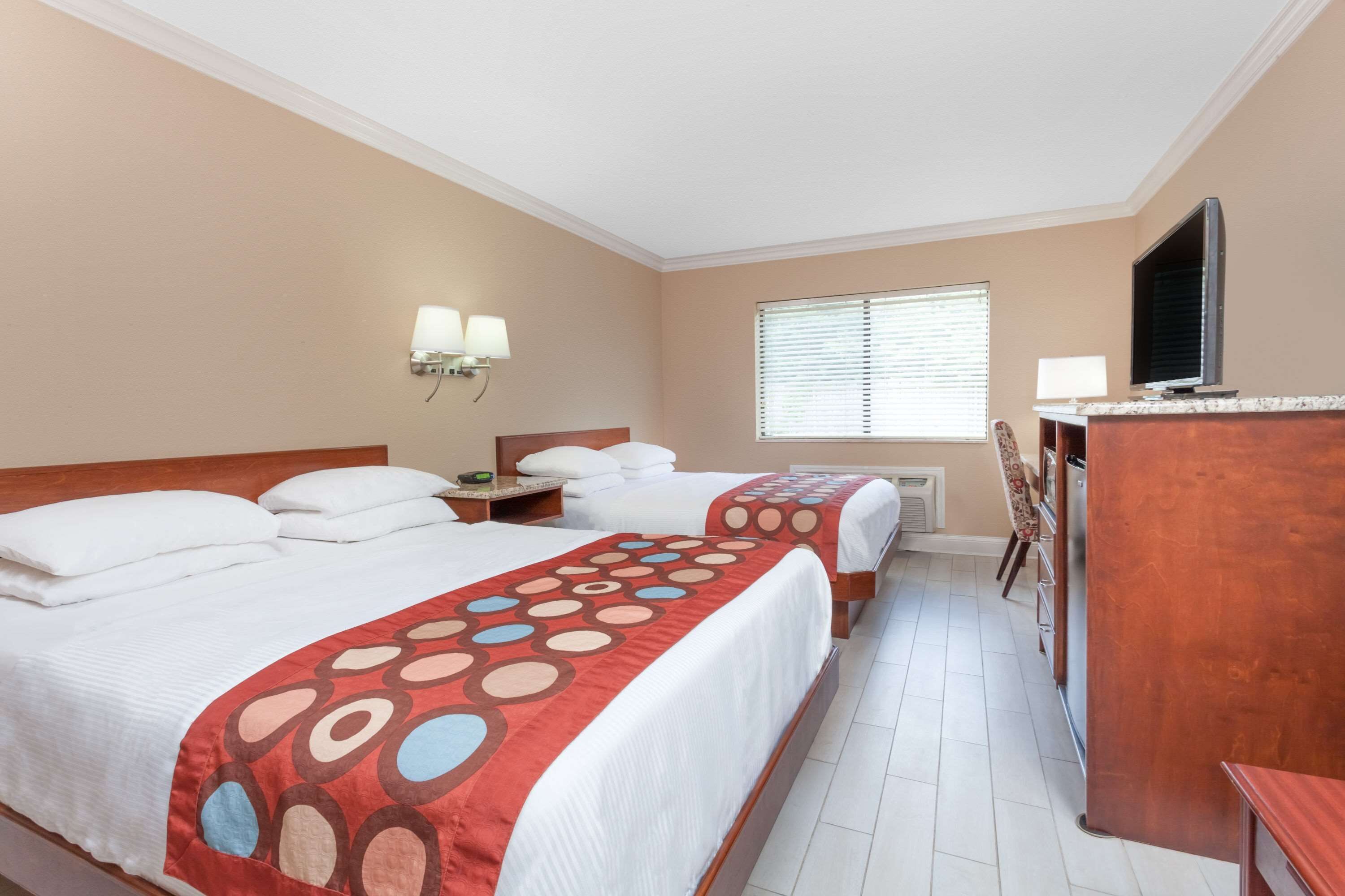 A hotel room with two beds, a desk, a TV, and a window with blinds.