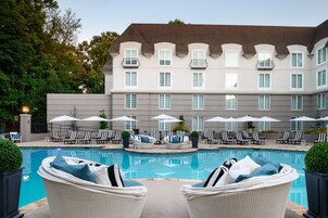Un hotel con piscina, asientos exteriores con sombrillas, y un edificio grande con múltiples ventanas y balcones.