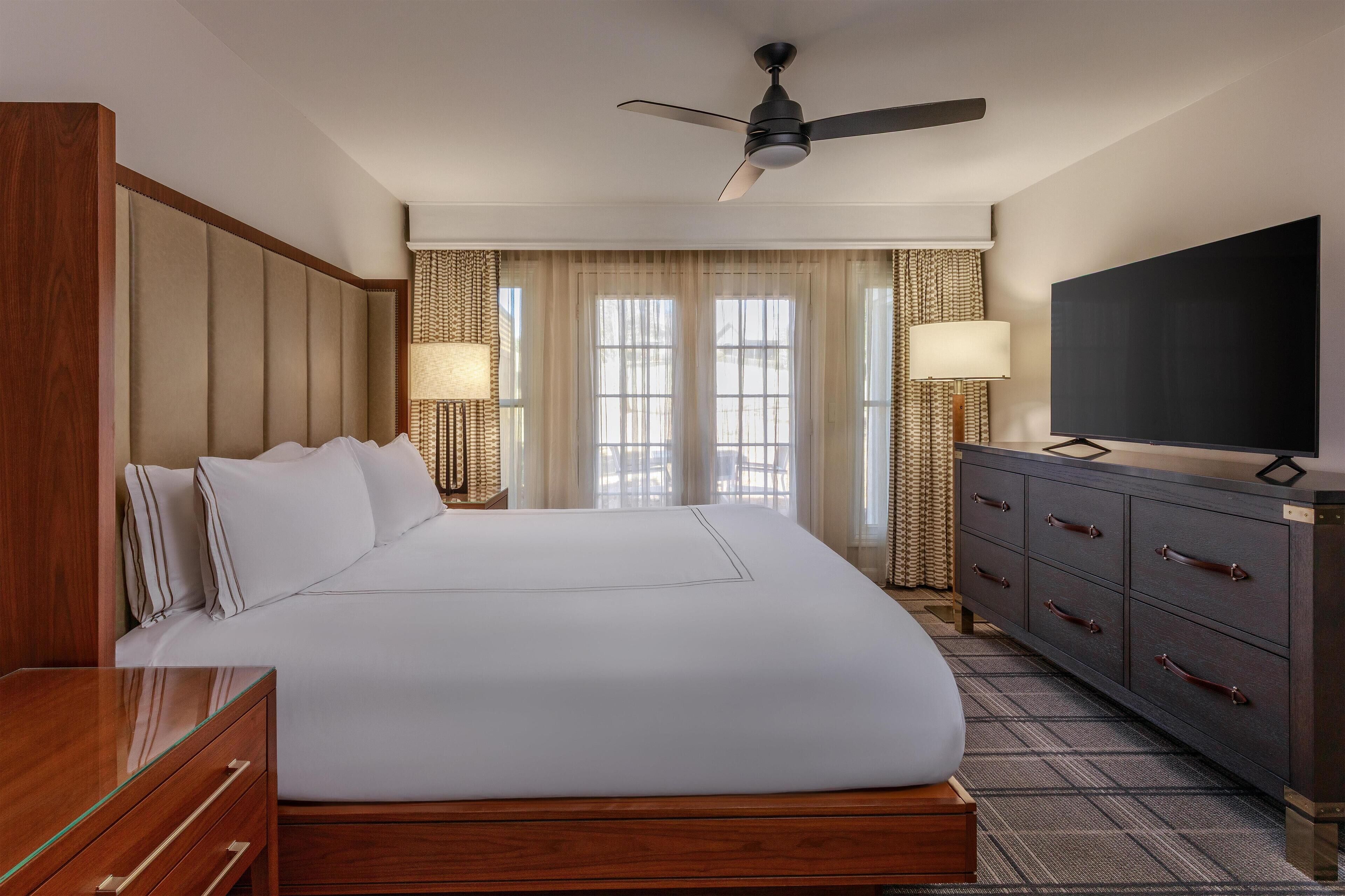 Ein Hotelzimmer mit einem großen Bett, einem Flachbildfernseher, einer Holzkommode und einem Deckenventilator.