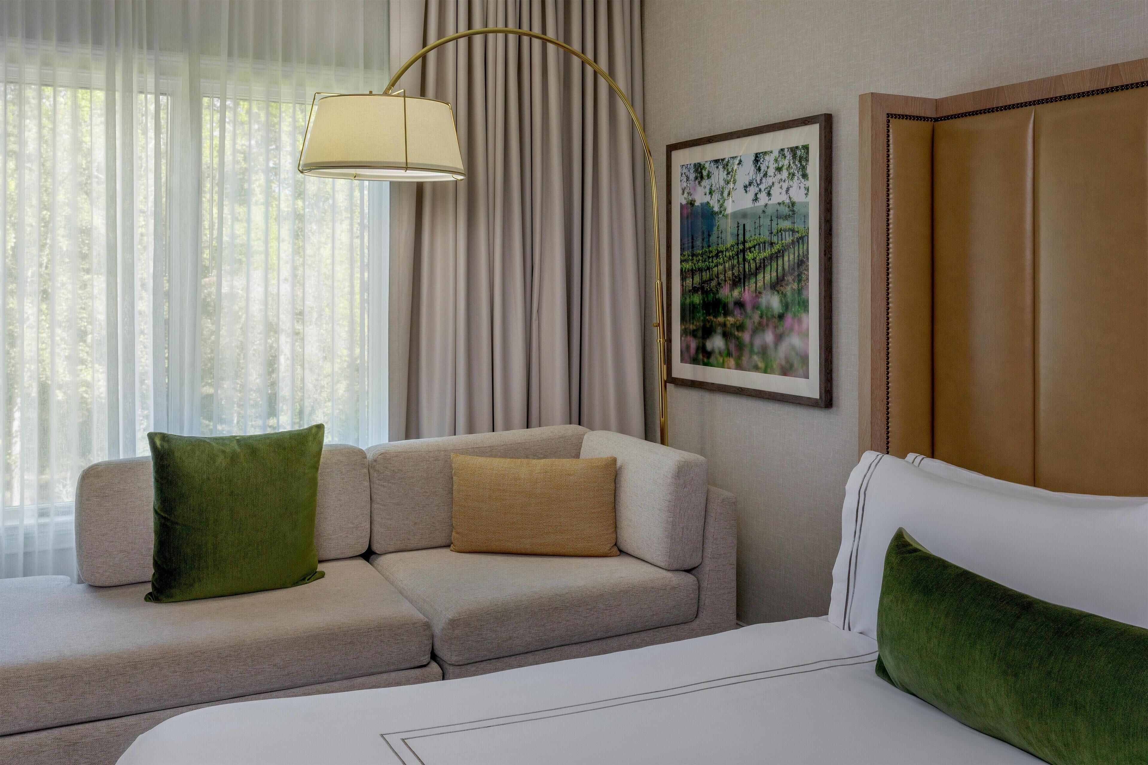 Una habitación de hotel con sofá, cama, lámpara, un cuadro y cortinas.