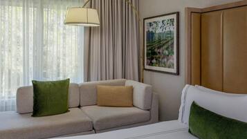 Una habitación de hotel con sofá, cama, lámpara, un cuadro y cortinas.
