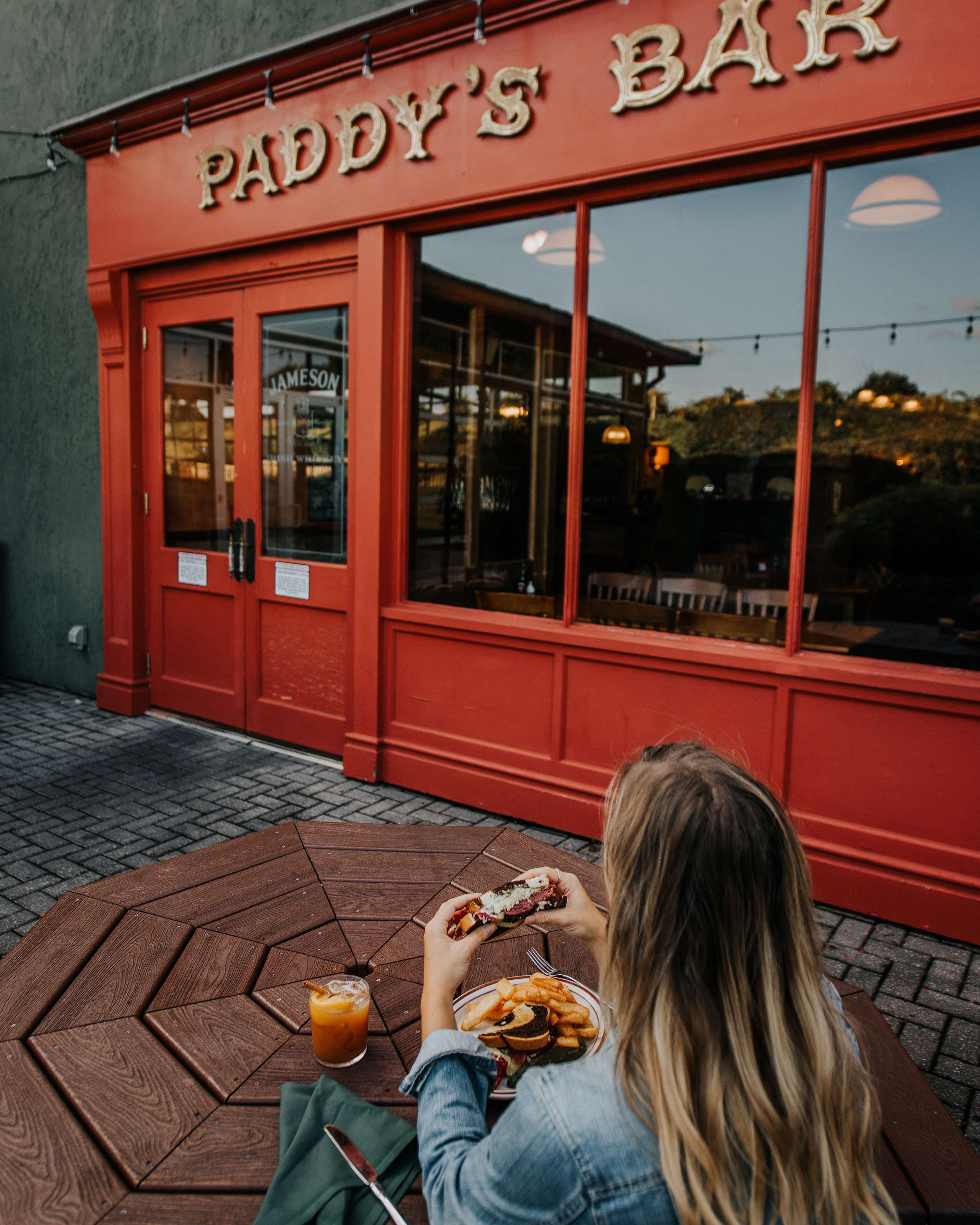 A person sitting at a wooden table outside a red building with a sign reading "PADDY'S BAR".