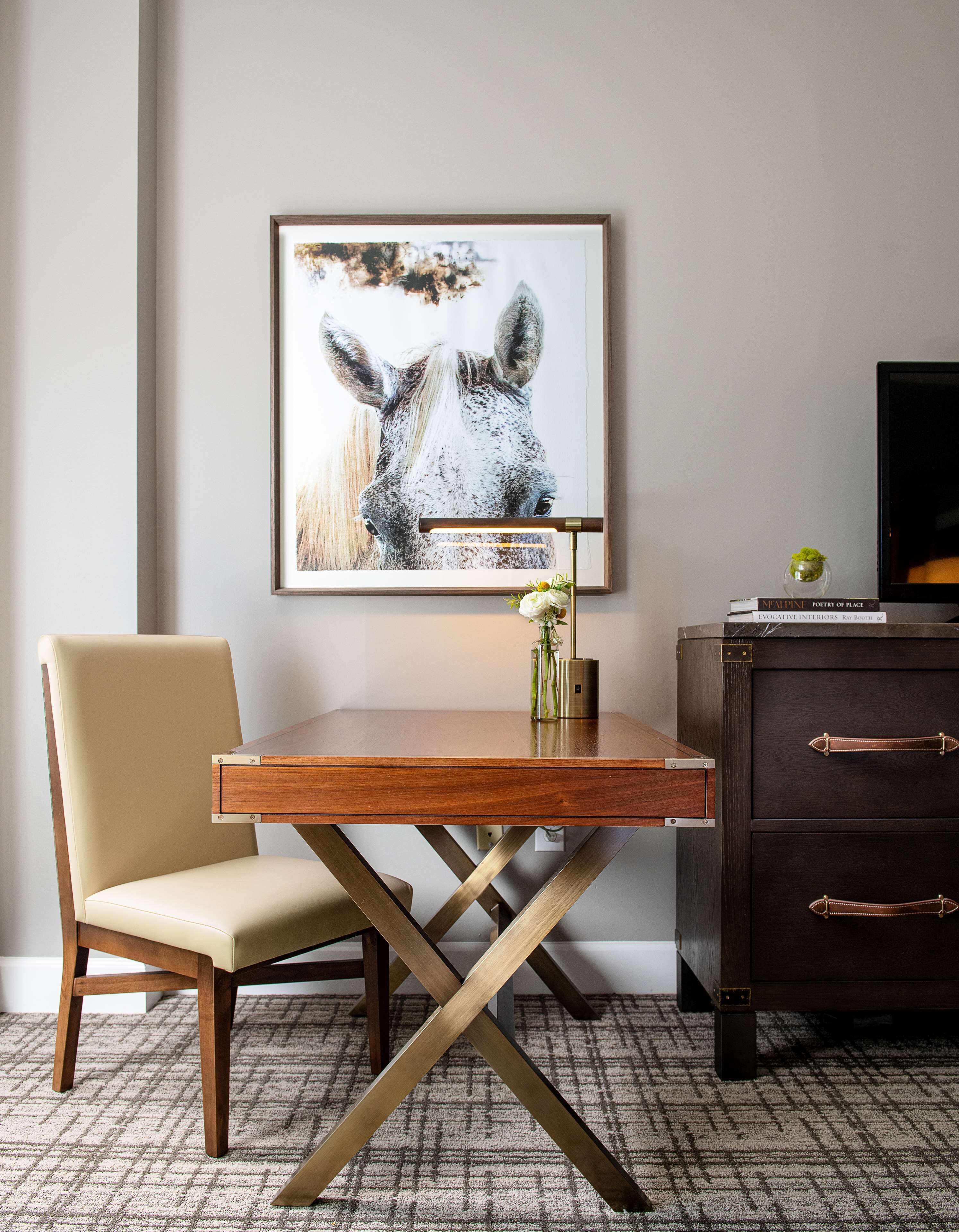 A modern room with a wooden desk, a chair, a framed artwork of a horse, a small vase with flowers, and a dark wooden dresser with drawers.