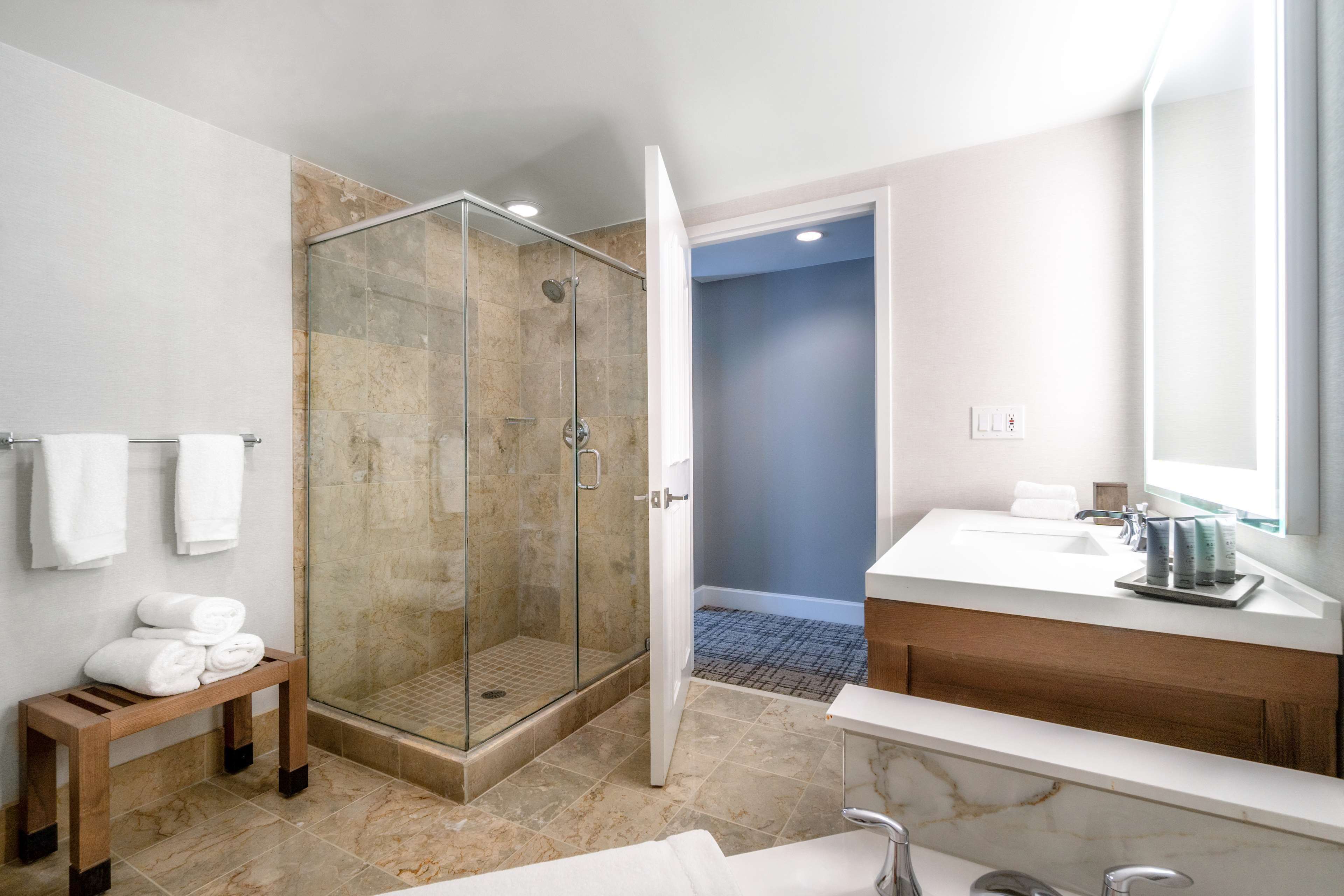 A modern bathroom with a glass-enclosed shower, a wooden bench with towels, a marble sink, and a large mirror.