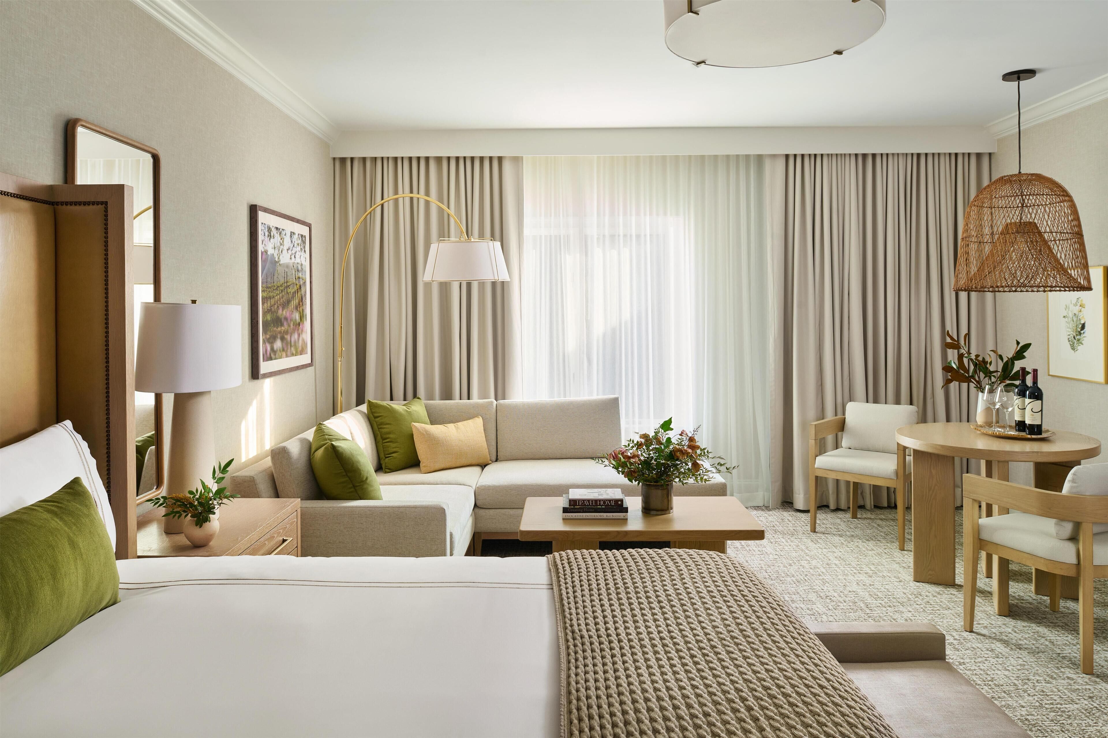 A hotel room with a large bed, a sofa, a coffee table, a side table with a lamp, a chair, a small table with a vase and a bottle, and a large window with curtains.