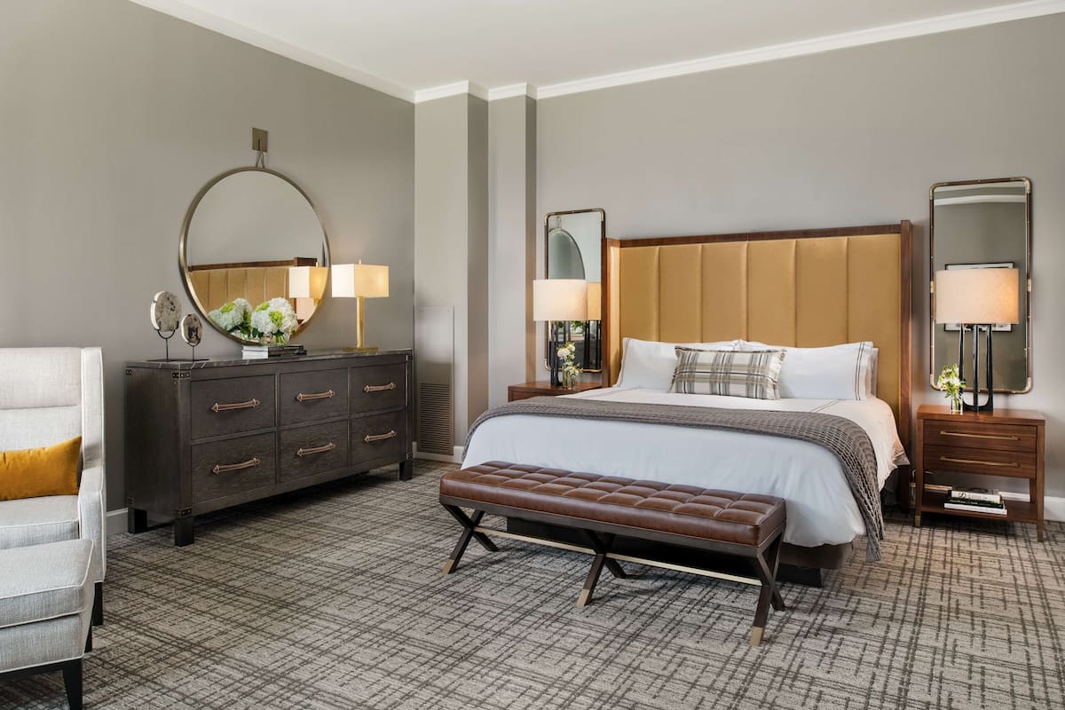 A hotel room with a large bed, a dresser, a bench, and two bedside tables with lamps.