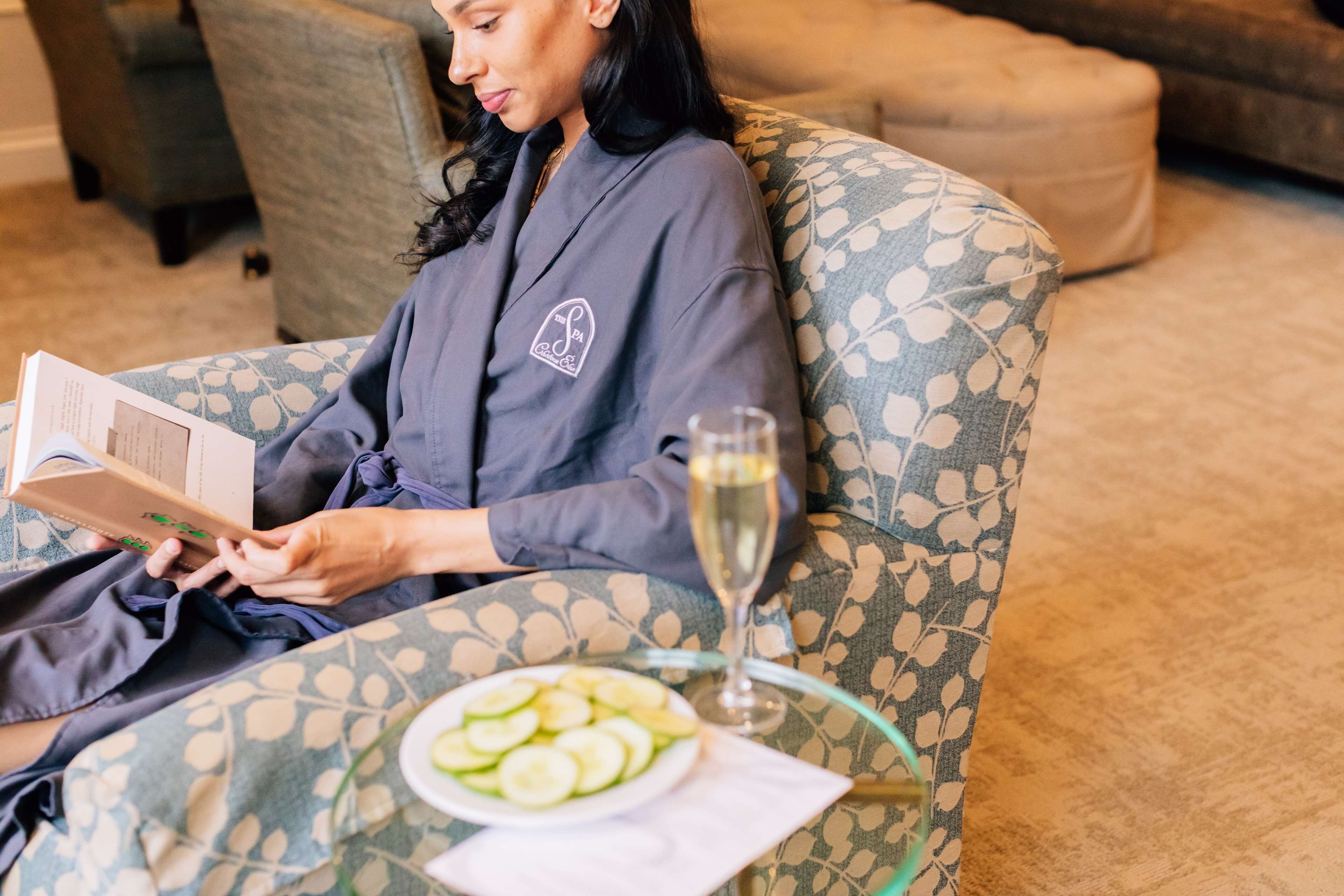 A person reading a book on a patterned sofa with a glass table beside them, containing a plate of sliced cucumbers and a glass of wine.