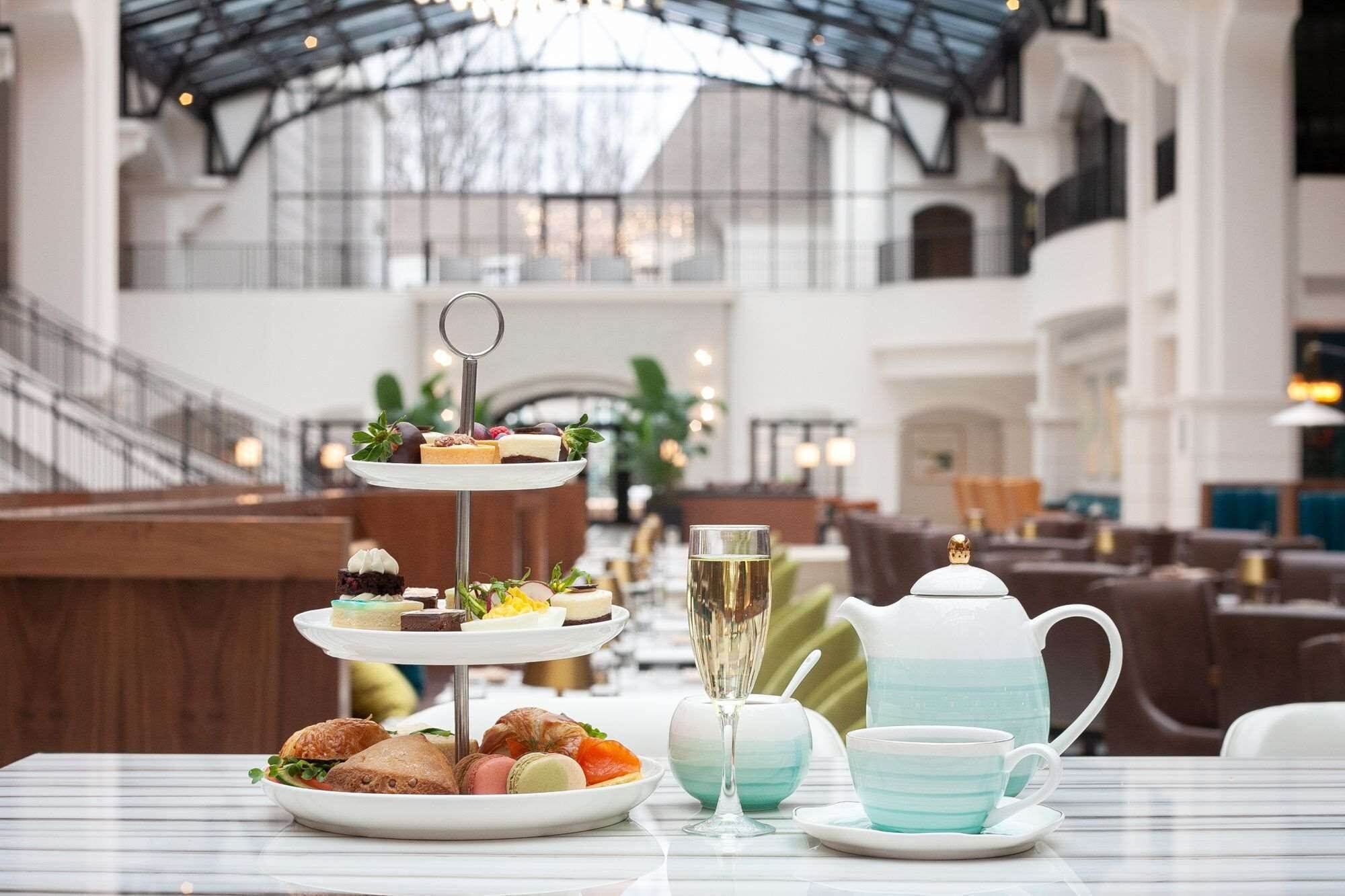 A three-tiered dessert stand with various pastries and a glass of champagne on a table in a spacious indoor area with high ceilings and seating in the background.