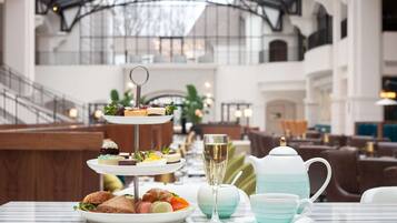 A three-tiered dessert stand with various pastries and a glass of champagne on a table in a spacious indoor area with high ceilings and seating in the background.