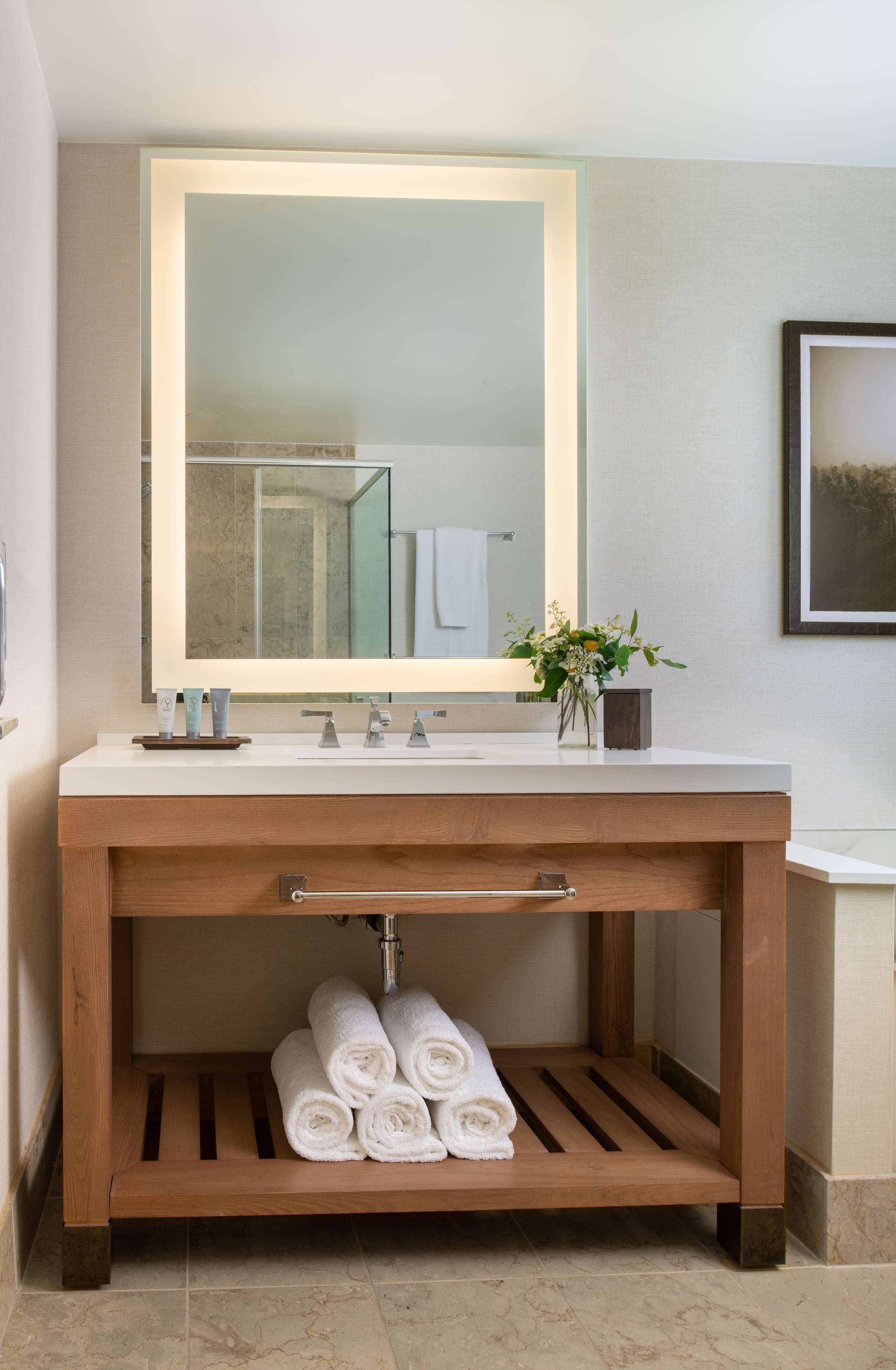 A bathroom with a wooden vanity, a large mirror, and a shelf with rolled towels.