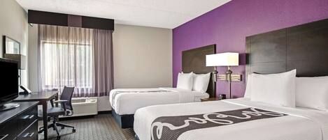 A hotel room with two beds, a desk, a TV, and a large window with curtains.