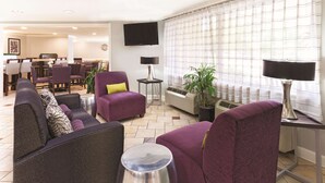Un hall d’hôtel avec des fauteuils violets, un coin repas, un téléviseur à écran plat et une grande fenêtre avec des rideaux.