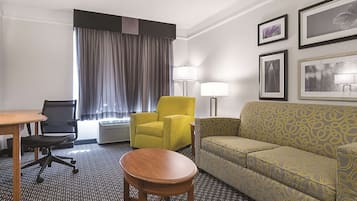 A hotel room with a desk, two chairs, a sofa, a small round table, a window with curtains, a floor lamp, and framed pictures on the wall.