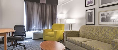 Habitación de hotel con escritorio, dos sillas, sofá, mesita redonda, ventana con cortinas, lámpara de pie y cuadros en la pared.