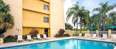 A hotel pool area with a clear blue pool, white lounge chairs, and palm trees.