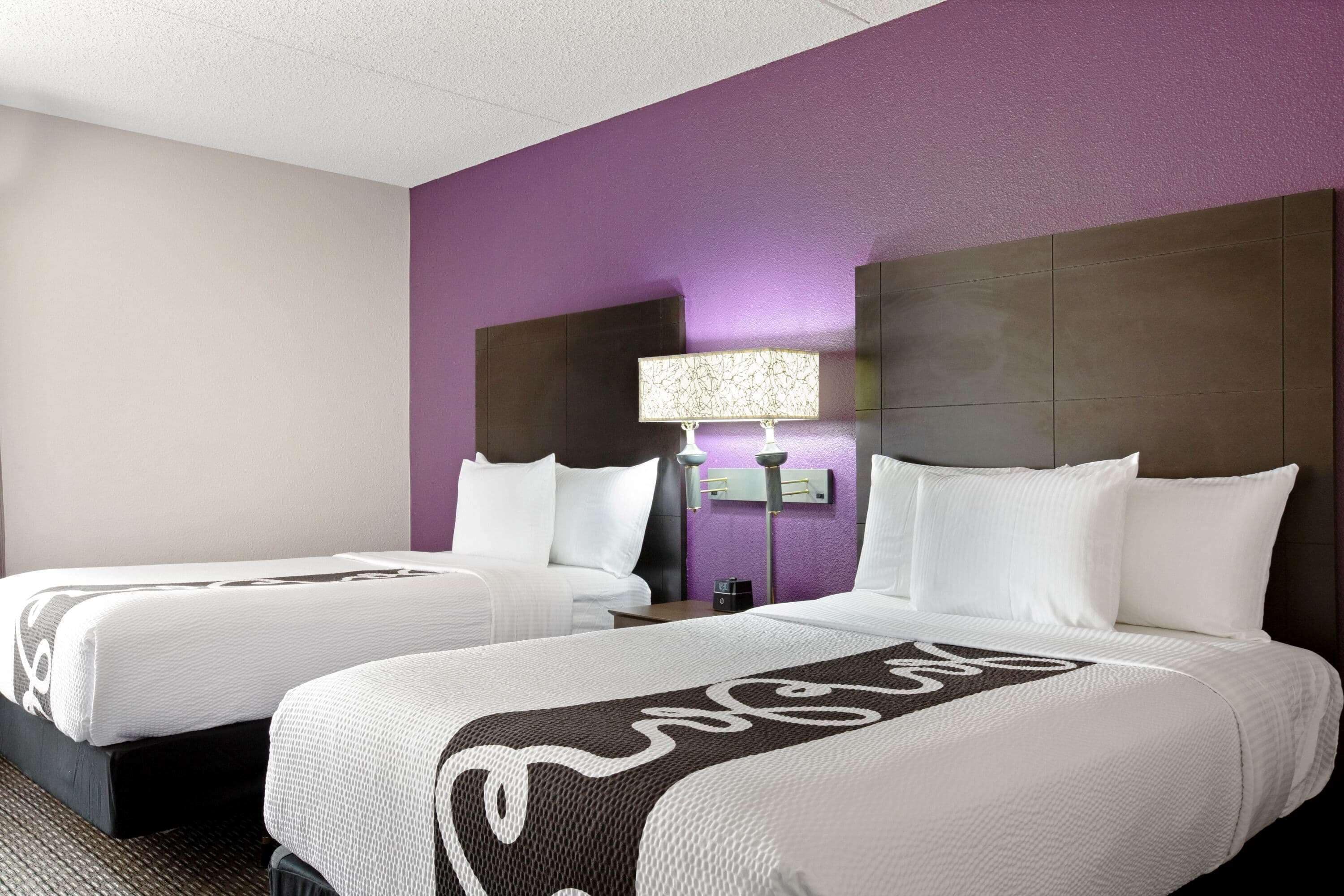 A hotel room with two beds, a purple accent wall, a nightstand with a lamp, and a headboard.