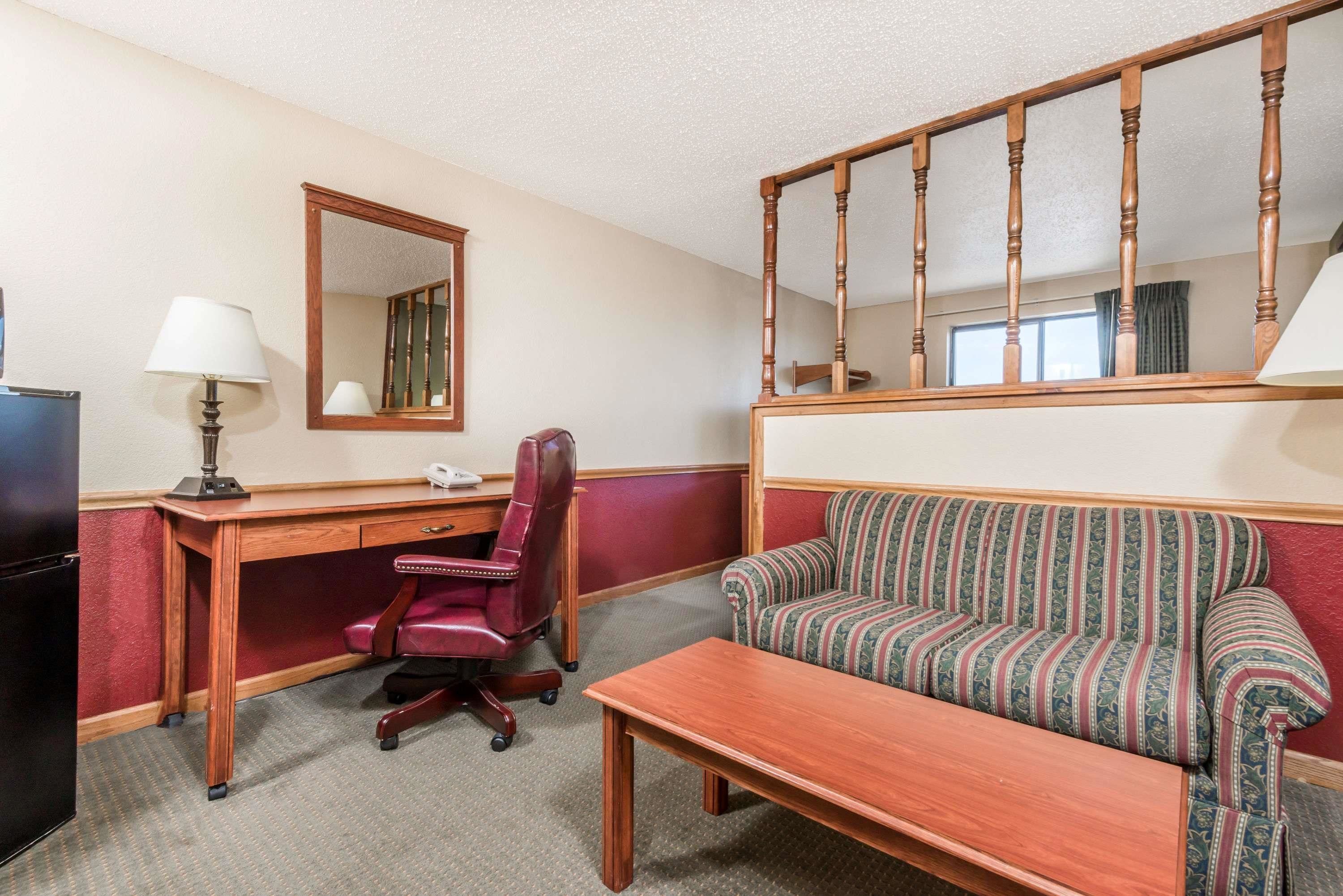 A hotel room with a wooden desk, a red office chair, a striped sofa, a wooden coffee table, and a mirror above the desk.