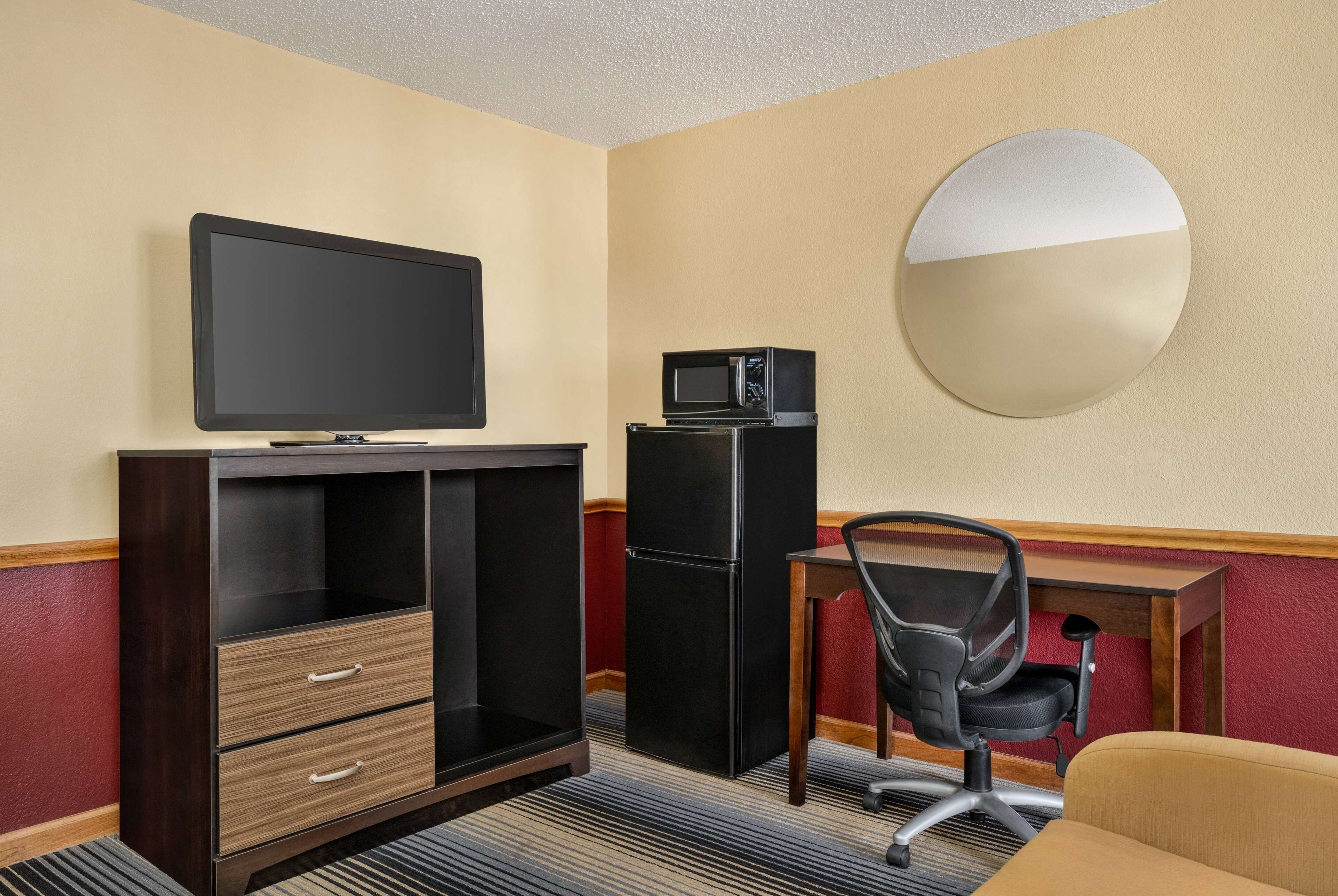 Habitación de hotel con televisión, microondas, refrigerador, escritorio, silla y espejo redondo en la pared.